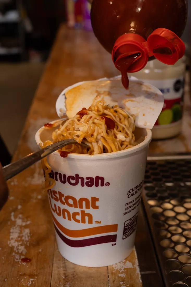 A cup of maruchas preparadas being finished with a squeeze of hot sauce
